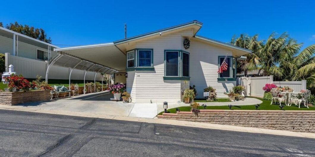 A beautifully maintained manufactured home in Encinitas featuring light exterior siding, tropical palm trees, manicured landscaping, a covered carport, and a private front patio in a peaceful senior living community.